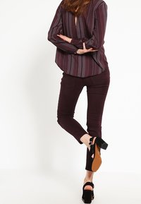 Vínové úzké kalhoty spojené s vzorovanou halenkou s dlouhými rukávy a výstřihem na zádech ve tvaru klíčové dírky. Modelka má na sobě černé sandály na platformě s pásky kolem kotníků.