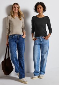 Två kvinnor står sida vid sida iförda långärmade skjortor, en beige och en svart, tillsammans med blå och ljusblå jeans.