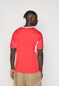 Piros sportmez fehér díszítésekkel és texturált ujjaikkal. Rövid ujjú, kerek nyakkivágású. A hátán a "suisse" felirat szerepel.