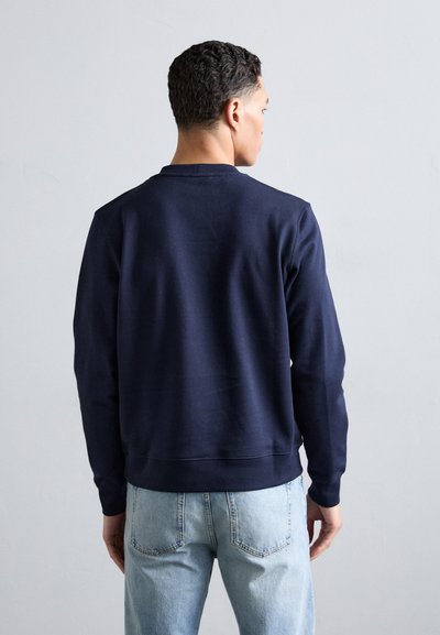 Felpa blu navy con maniche lunghe e polsini a coste, caratterizzata da un semplice colletto girocollo e una texture morbida, abbinata a jeans blu chiaro.