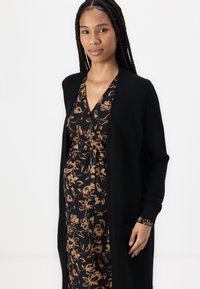 Cardigan nero lavorato a maglia con apertura frontale, maniche lunghe e tasche, indossato sopra un vestito nero con un motivo floreale marrone.