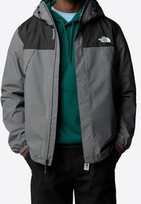 Homme portant une veste North Face grise et noire sur un sweat à capuche vert avec les mains dans les poches d'un pantalon noir.
