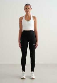 Fehér sport crop top logóval, magas derekú fekete leggingsszel, logó részletekkel. A modellen fehér sportcipők vannak. Egyszínű, testhezálló dizájn.