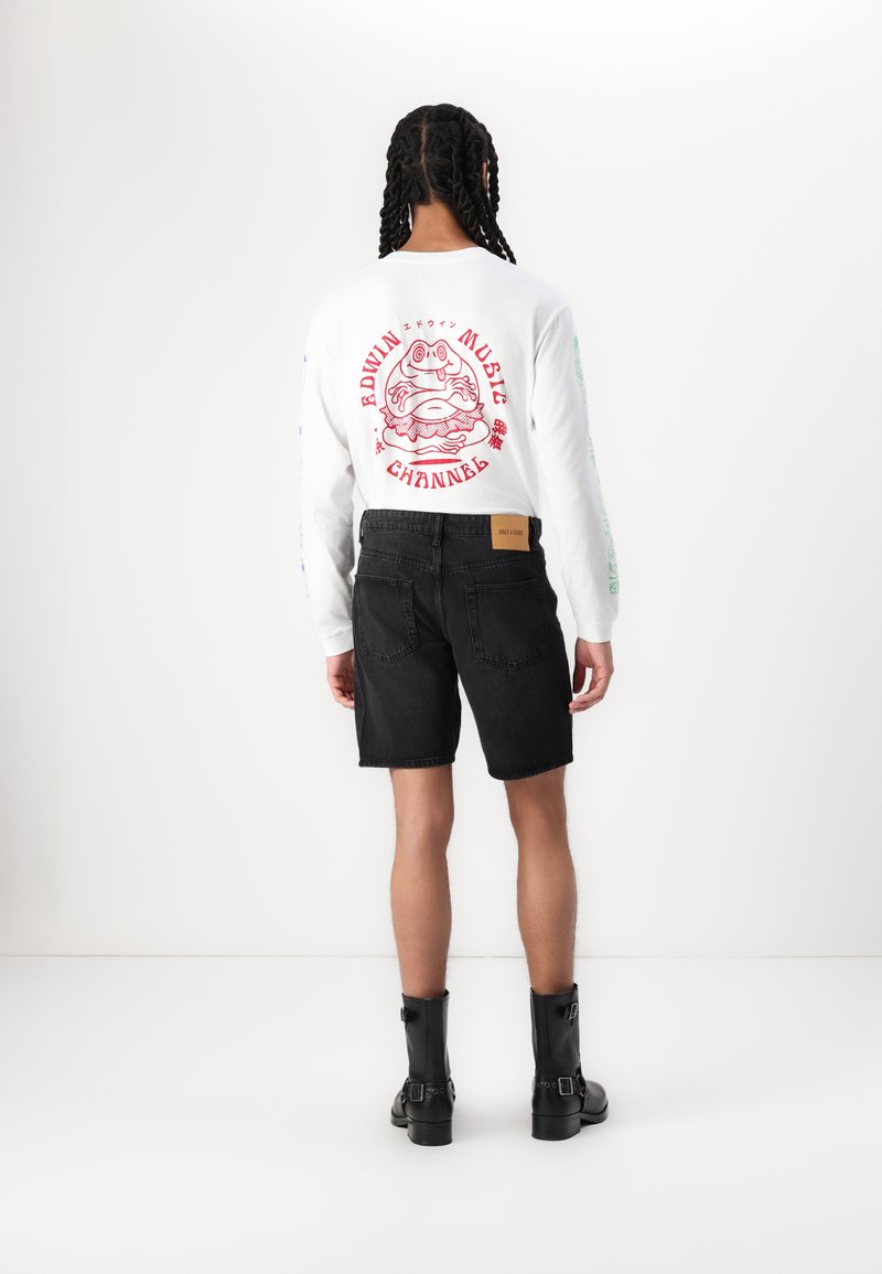 Hvid langærmet skjorte med rød grafik, sort denim shorts og sorte ankelstøvler. Skjorten har farverig tekst langs ærmerne.
