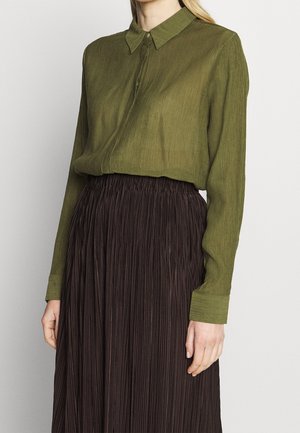 Kobieta w oliwkowej, długiej bluzce z kołnierzem, wsuniętej w spódnicę plisowaną o wysokim stanie w ciemnobrązowym kolorze.