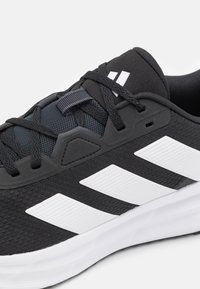 Bottines de sport noires avec une tige texturée comportant des sections en maille, trois rayures blanches et une semelle en caoutchouc blanche. Les lacets sont noirs.