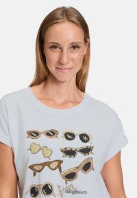 T-shirt blanc ornée de plusieurs lunettes de soleil illustrées en or, noir et brun. Manches courtes, col rond, design décontracté.