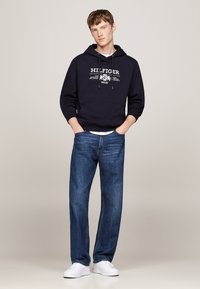 Sudadera con capucha de color navy con impresión de logo blanco, combinada con jeans de mezclilla azules y zapatillas blancas. Corte casual con bolsillo tipo canguro en la parte frontal.
