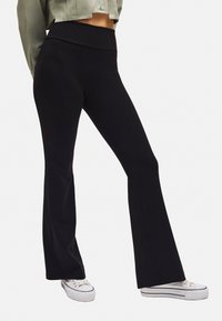 Leggings negros acampanados hechos de un tejido elástico con una cinturilla alta. Combinados con zapatillas blancas. Presentan una textura suave y líneas limpias.