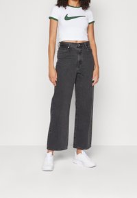 Mustat leveälahkeiset farkut korkealla vyötäröllä, yhdistetty valkoiseen lyhytpaitaiseen t-paitaan, jossa on vihreä Nike-logo, sekä valkoisiin urheilukenkäisiin.