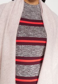 Frau, die einen kastanienbraunen und grauen gestreiften Strickpullover mit leuchtend roten Streifen trägt, darunter einen weichen, hellrosa Schal oder Cardigan.