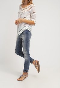 Femme portant un haut en crochet blanc, un jean déchiré raccourci et des sandales plates à lanières, se tenant avec une jambe légèrement levée.