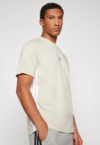 Hellbeige T-Shirt mit kurzen Ärmeln, das ein kleines lila Logo auf der Brust und weiße Seitenstreifen aufweist. Aus weichem Baumwollmaterial.