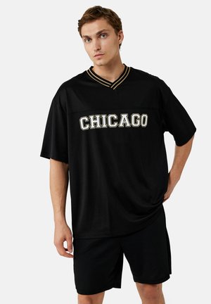 Ung mand med kort hår iført en løs sort skjorte med teksten "CHICAGO" og matchende sorte shorts, stående mod en ensfarvet baggrund.