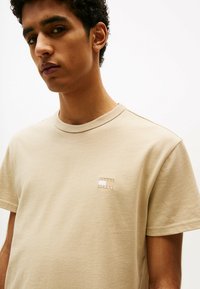 T-shirt beige a maniche corte realizzato in tessuto morbido, con una piccola patch logo ricamata sul lato sinistro del petto, scollatura rotonda e vestibilità rilassata.