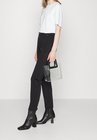 Černé tapered kalhoty, bílé tričko s krátkým rukávem a černé kotníkové boty. Průhledná plastová taška s černou rukojetí. Hladké textury a minimalistický design.