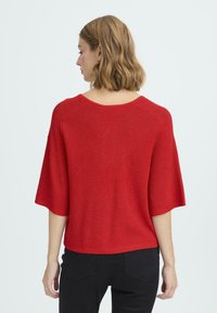 Crvena pletena majica s kratkim, širokim rukavima i lagano izrezanim dekolteom. Teksturirani materijal s opuštenim krojem, uparen s tamnim hlačama.