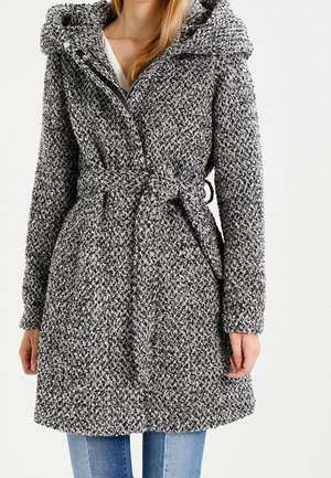 Kobieta w teksturowanym, szarym wełnianym płaszczu z kapturem, paskiem w talii i bocznymi kieszeniami, sięgającym do kolan, na jasnoniebieskich dżinsach.