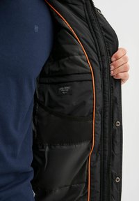 Sort polstret jakke med orange detaljer, med lynlåslukning, indvendige lommer og en sort mesh-lomme, båret over en blå skjorte.