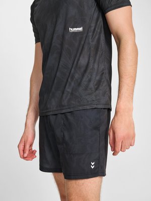 Mężczyzna w czarnej sportowej koszulce i szortach hummel Performance, stoi z ramionami swobodnie opuszczonymi wzdłuż ciała, na jednolitym tle.