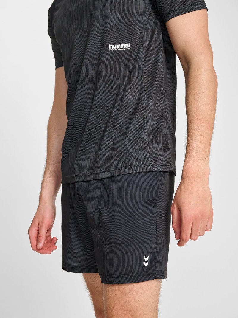 Mann trägt schwarzes hummel Performance Sportshirt und Shorts, steht mit entspannten Armen seitlich am Körper vor einfarbigem Hintergrund.