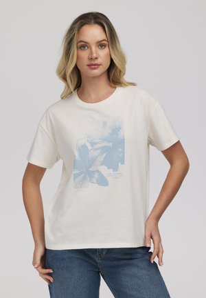 Kobieta o blond włosach do ramion, ubrana w biały t-shirt z kwiatowym wzorem i niebieskie dżinsy, stojąca z jedną ręką na biodrze.