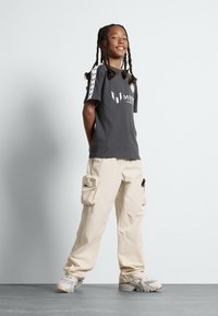 Camiseta gris de manga corta con logo y acentos estampados, combinada con pantalones cargo beige con bolsillos. El calzado es de color gris claro.