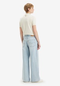 Lys beige polo skjorte med korte ermer, kombinert med vide lyseblå denimjeans med baklommer og et logomerke.