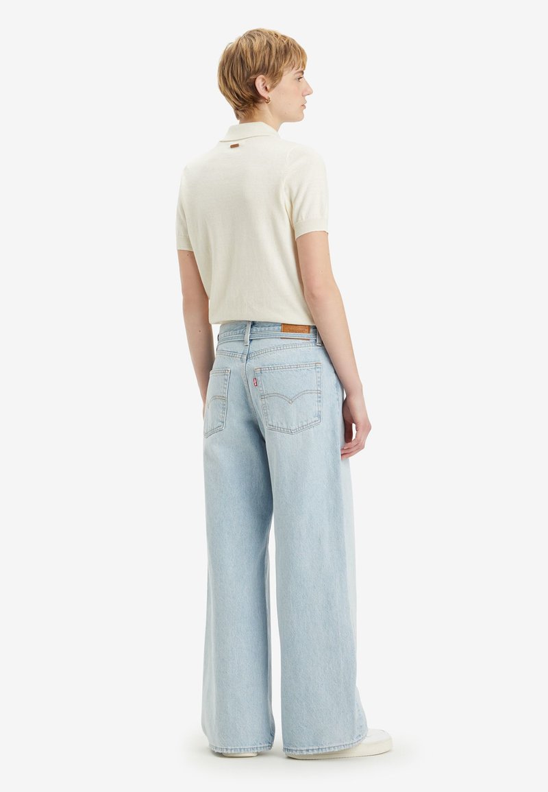 Polo shirt beige clair à manches courtes, associé à un jean en denim bleu clair à jambes larges avec poches arrière et une étiquette de logo.