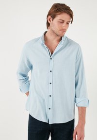 Camisa de botones de manga larga en un tejido ligero de color azul claro; cuenta con un cuello, botones negros y puños doblados.
