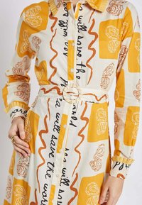 Button-up dress made of lightweight fabric. Features yellow-orange and white patterns with text and a textured collar. Includes a circular belt buckle.