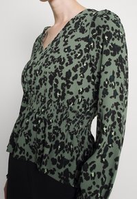 Grüne Bluse mit schwarzem Leopardenmuster, V-Ausschnitt, langen puffärmeligen Ärmeln, gerafftem Bund und geschichtetem Rüschensaum. Glatte Stofftextur.