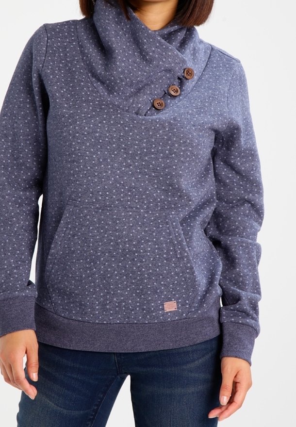 Femme portant un pull à capuche bleu à pois avec trois boutons en bois sur le col cheminée et une poche kangourou devant, associé à un jean foncé.
