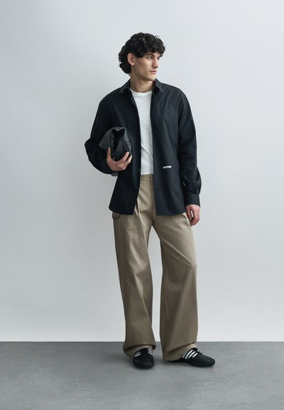 Ung mand iført sort skjorte, hvid T-shirt, beige bukser med vide ben og sorte sneakers, der holder en foldet sort læderclutch, stående foran en grå væg.