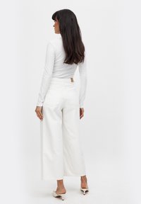 Witte blouse met lange mouwen, gecombineerd met hooggeknoopte, wijdvallende witte jeans. Opvallende details zijn achterzakken en een tan leren label aan de taille.