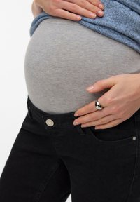 Έγκυος που φοράει μαύρο τζιν εγκυμοσύνης και γκρι μπλούζα, κρατώντας απαλά την κοιλιά της με το ένα χέρι και ακουμπώντας το άλλο στη μέση του παντελονιού.