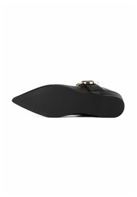Chaussure plate noire à bout pointu avec un détail de boucle dorée vue du côté de la semelle sur fond blanc.