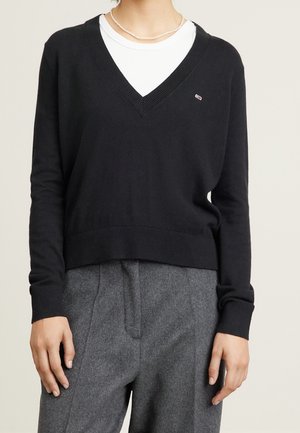 Personne portant un pull noir col en V par-dessus une chemise blanche, un pantalon gris taille haute et un court collier de perles, debout devant un fond uni.