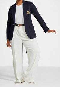 Navyblazer med guldlogotyp, vit t-shirt, vita vida byxor och en brunrandig bälte, med sportiga sneakers.