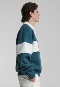 Sweatshirt în dungi turcoaz și alb, cu guler ribbit, material texturat și croială relaxată. Include un logo vizibil și un design casual.