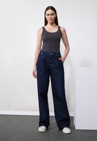 Jeans ampi di colore blu scuro con vita alta, tasche frontali e chiusura con bottoni, abbinati a un top aderente grigio e sneakers bianche.