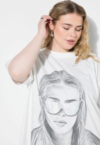 Bílé oversized tričko s podrobnou grafikou ženy v reflexních slunečních brýlích. Měkká tkanina s volným střihem a krátkými rukávy.