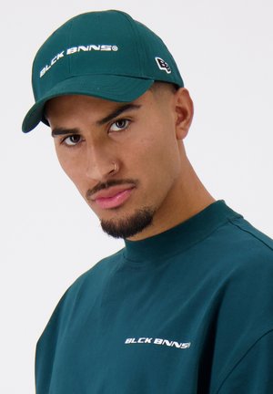 Groene baseballpet met het logo "BLCK BNNS" in het wit, plat vizier ontwerp. Wordt gedragen met een bijpassend groen shirt met ronde hals waarin hetzelfde logo staat.