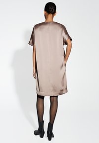 Satinbraunes Kleid mit kurzen Ärmeln, lockerer Passform, seitlichen Taschen und geradem Saum, kombiniert mit schwarzen Knöchelstiefeln und transparenter schwarzer Strumpfhose.