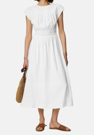 Woman wearing a white midi dress with gathered waist and short sleeves, holding a straw tote bag and wearing brown slide sandals.