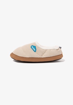 Zapatilla beige acolchada con suela marrón, forro interior de felpa blanca y parche azul con logo en el lateral, diseñada para la comodidad en interiores.