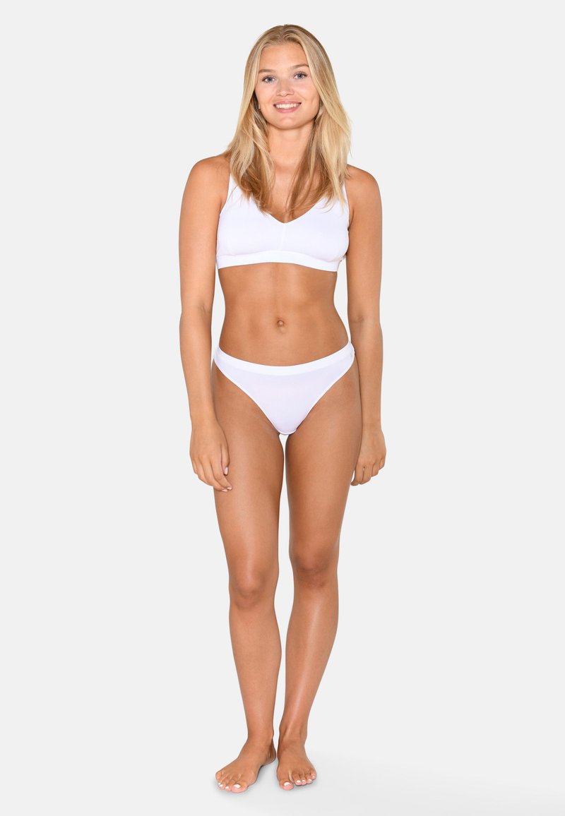 Jeune femme debout pieds nus, portant un bikini blanc, souriante, aux longs cheveux blonds sur un fond gris clair uni.