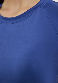 Top sportiv albastru cu guler rotund, textură netedă și mâneci raglan, subliniind un design ajustat cu detalii de cusătură.