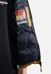Giacca navy con una texture trapuntata, caratterizzata da un patch logo colorato con montagne e sole, e un accento dorato lungo la cerniera.