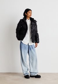 Musta untuvatakki kiiltävällä pinnalla ja vetoketjullisilla taskuilla, valkoisen napillisen paidan ja vaaleansiniset leveät farkut päällä.